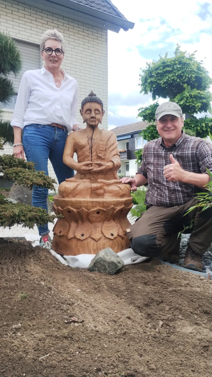 Zwei Personen stehen neben einer Buddha-Statue im Garten, einer gibt Daumen hoch.